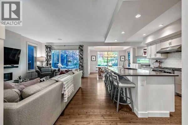 Open-concept living area with a grey sectional sofa, kitchen island with bar stools, and dining area. - 21 Ascot Crescent Sw, Calgary