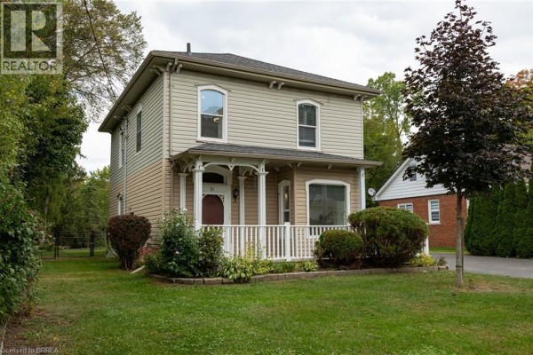 Side view of a two-story beige house with a covered front porch and lush greenery. - 21 Mccall Street, Simcoe