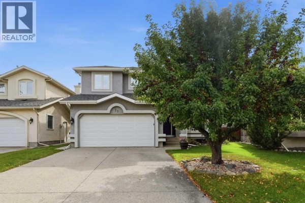 Front view of a two-story grey house with a large double garage door and a mature tree in the yard. - 21 Mt Robson Close Se, Calgary