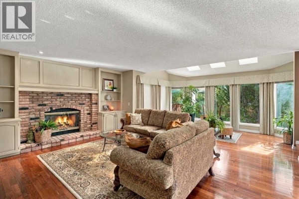 Living room featuring a brick fireplace, built-in shelves, hardwood floors, and large windows with greenery views. - 21 Mt Robson Close Se, Calgary