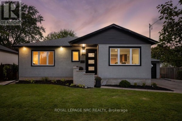 Side view of the grey stucco bungalow with a black garage door and brick chimney. - 21 Silverdale Drive, St  Catharines