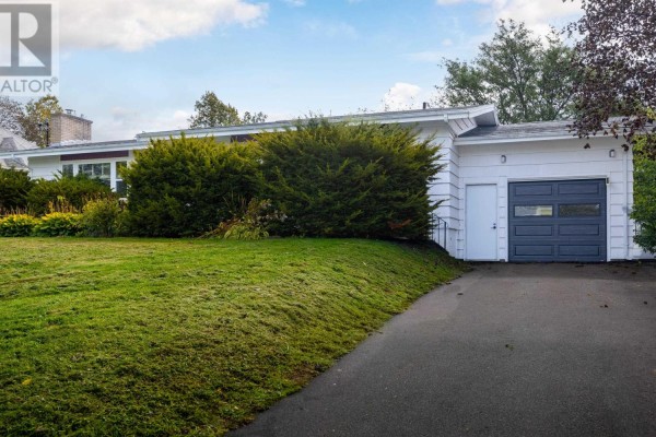 Side view of a white house with a dark grey garage door and a sloping green lawn. - 21 Smallwood Crescent, Charlottetown
