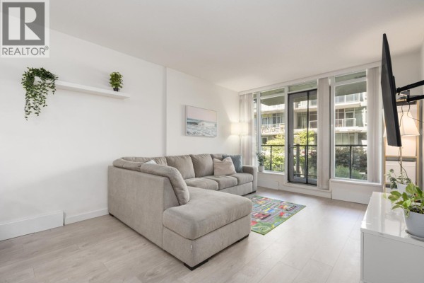 A bright living area with a sectional sofa, white walls, and a large window. - 210 3168 Riverwalk Avenue, Vancouver