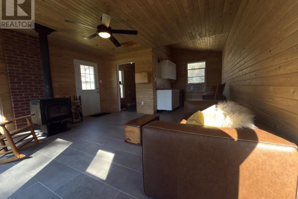 Living area with wood-paneled walls, a brown sofa, wood stove, and a small kitchenette. - 210 C Storms Road, Prince Edward County