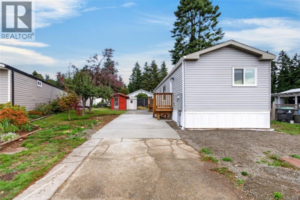 View of the mobile home's exterior, driveway, and surrounding lot with trees and other mobile homes. - 2104 Pemberton Pl, Comox