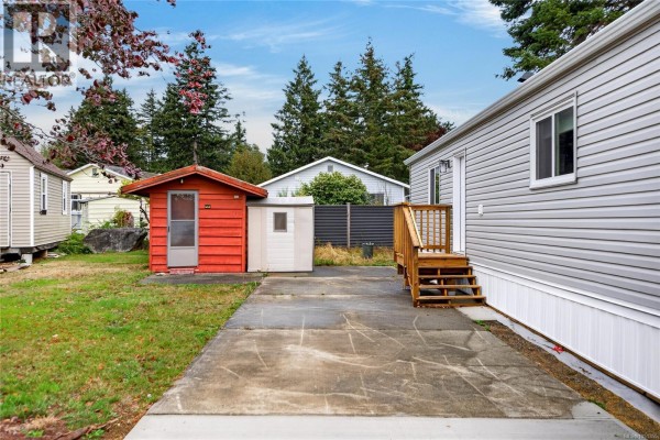 Backyard of the mobile home featuring a concrete patio, red shed, and storage unit. - 2104 Pemberton Pl, Comox