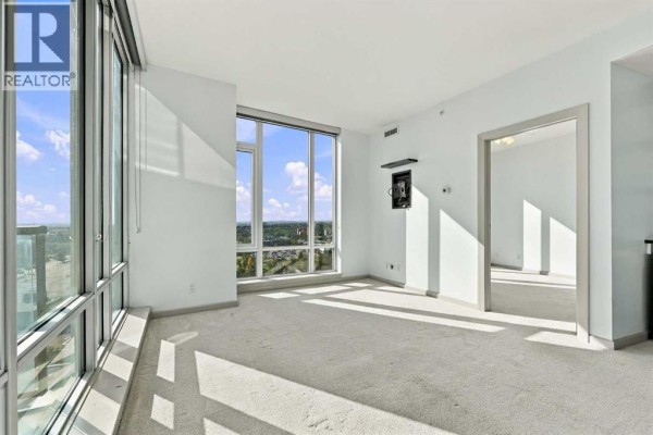 Empty living area with beige carpet, large windows, and doorway to another room. - 2106, 210 15 Avenue Se, Calgary