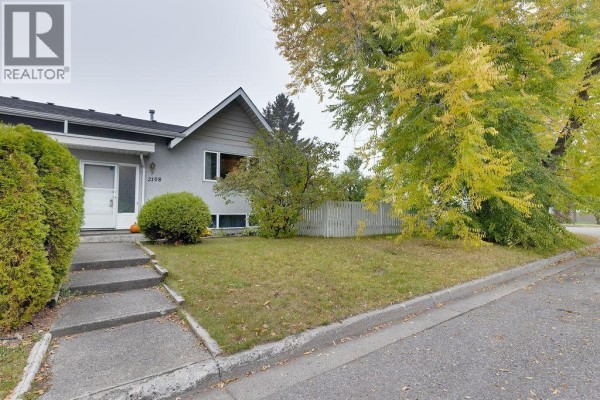 Side exterior view of a duplex with a white picket fence and autumn trees. - 2108 6th Avenue, Prince George