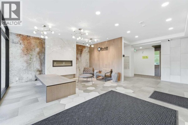 Apartment building lobby with a reception desk, elevator, seating area, and textured walls. - 211, 19661 40 Street Se, Calgary