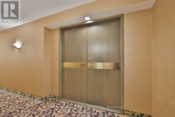 Apartment entrance door labeled '211' with wood finish and brass accents on textured walls. - 211 - 1800 The Collegeway, Mississauga