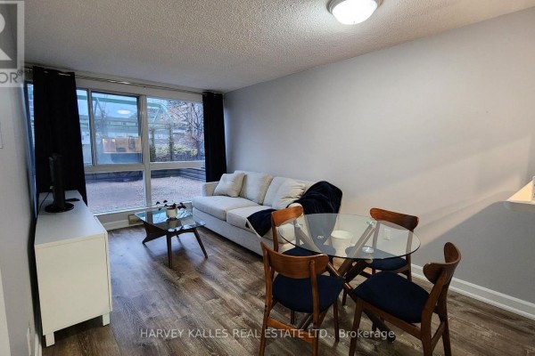 Living area with sofa, dining set, TV console, and large window overlooking a courtyard. - 211 - 7 Broadway Avenue, Toronto