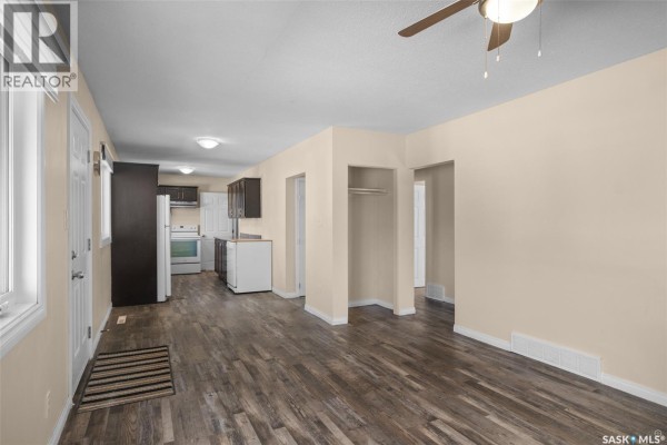 Open concept living area showing beige walls, wood-look vinyl plank flooring, a kitchen with dark cabinets, and a hallway. - 211 31st Street E, Prince Albert