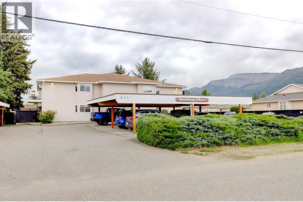 Exterior view of a beige apartment building with a covered carport and mountain backdrop. - 211 Kildonan Avenue Unit# 212, Enderby