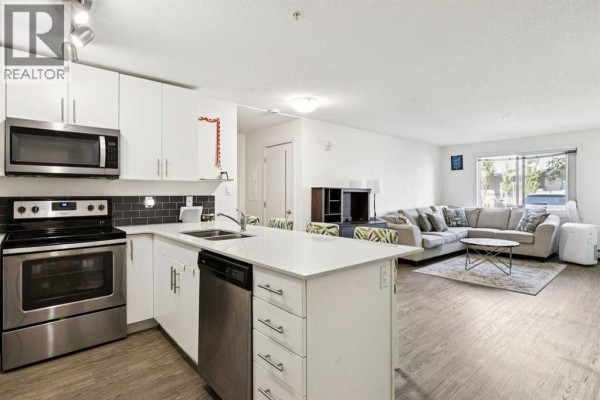 Open concept living and kitchen area with white cabinets, stainless steel appliances, sectional sofa, and coffee table. - 2115, 181 Skyview Ranch Manor Ne, Calgary