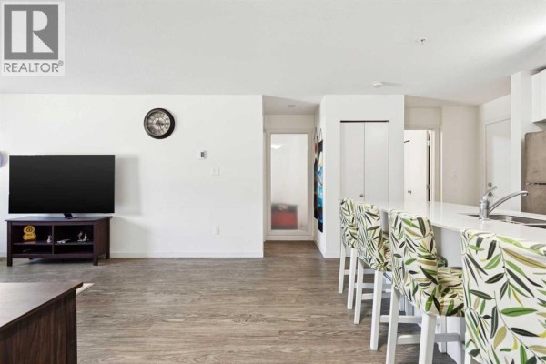 Living room with a large TV on a dark wood stand, light wood laminate flooring, and view into the kitchen with bar stools. - 2115, 181 Skyview Ranch Manor Ne, Calgary