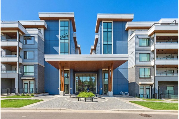 Building entrance with large glass doors, blue siding, and wooden architectural features. - 212 6220 Gooseberry Drive, Surrey