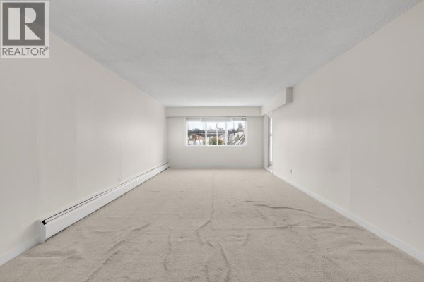Empty living room with beige carpet, white walls, and a large window. - 212 6420 Buswell Street, Richmond