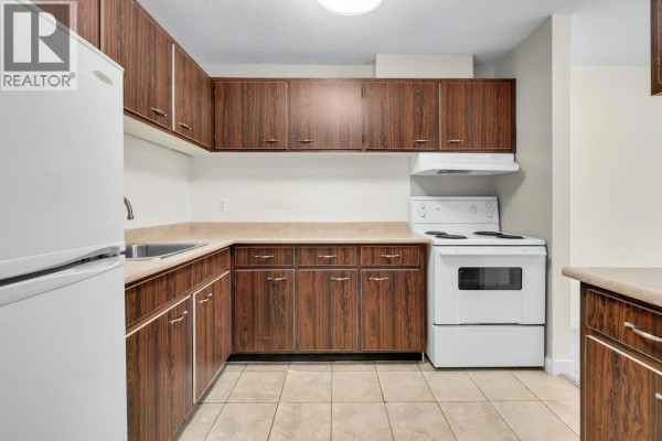 Living area with beige carpet, white walls, and view into a small kitchen area. - 212 6420 Buswell Street, Richmond