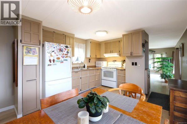 Kitchen with tan cabinets, white appliances, a wooden table with placemats, and a view into a living area. - 2129 Rama Rd, Campbell River