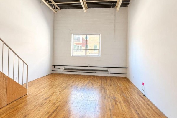 Living area with white brick walls, hardwood floors, a window overlooking a building, and a heating unit. - 213 - 227 Sterling Road E, Toronto