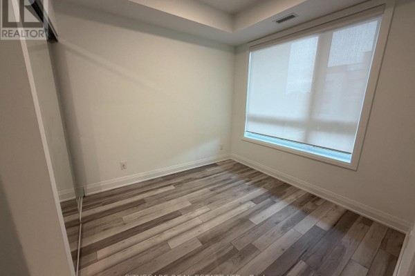 Empty bedroom with grey wood-look flooring, white walls, and large roller-blinded window. - 213 - 3250 Carding Mill Trail, Oakville