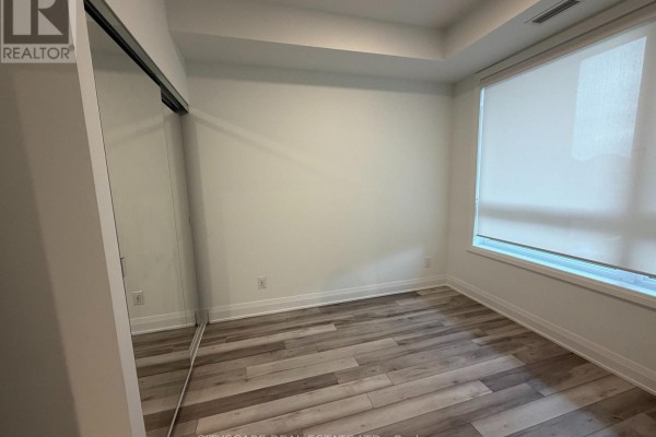 Empty bedroom with grey wood-look flooring, white walls, and large roller-blinded window. - 213 - 3250 Carding Mill Trail, Oakville