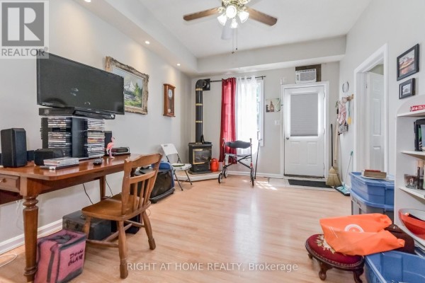 Living area with light wood floors, a wood-burning stove, desk with chair, and a television on a shelf. - 213 Main Street E, Minto