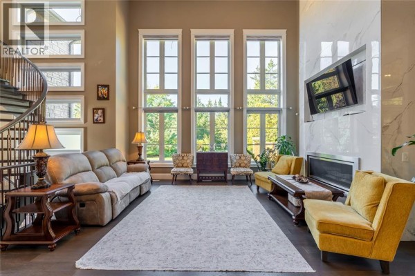 Living room with a large rug, beige sofa, two yellow chairs, and a wall of tall windows overlooking trees. - 2132 Huron Shores Drive, Sarnia