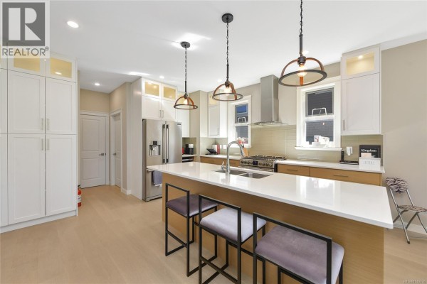 Modern kitchen with white cabinets, stainless steel appliances, and a large island with pendant lights. - 2136 Champions Way, Langford