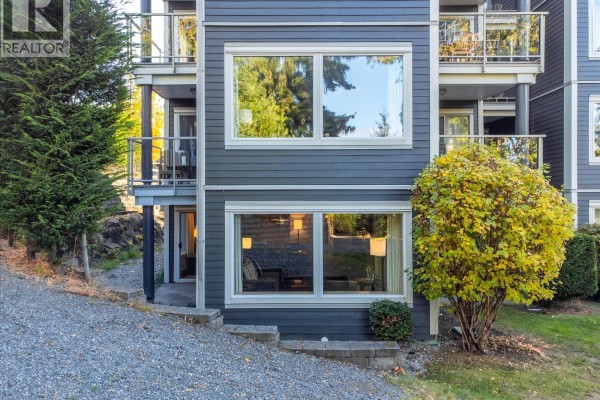 Apartment building exterior with dark grey siding, large windows, and green foliage. - 214 1600 Stroulger Rd, Nanoose Bay