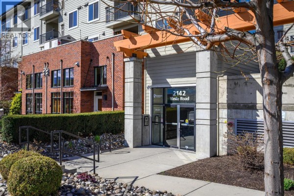 Main entrance of the building featuring glass doors, stone pathway, and decorative landscaping. - 2142 Vasile Road Unit# 211, Kelowna