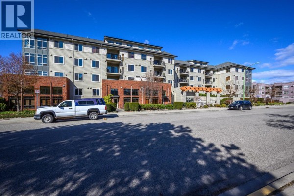 Street view of the apartment complex with vehicles parked and surrounding landscaping. - 2142 Vasile Road Unit# 211, Kelowna