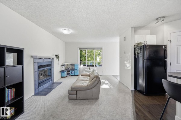 Living room with a fireplace, beige couch, window view, black refrigerator, and carpet flooring. - 215 85 St Sw, Edmonton