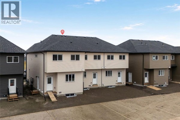 Rear exterior view of townhouses with beige siding, dark roofs, and small backyard patios. - 21527 Sheriff King Street Sw, Calgary