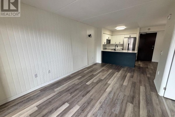 Living area showing wood-look flooring, white paneling, and a view of the kitchen and entrance. - 216 - 40 Via Rosedale Avenue, Brampton