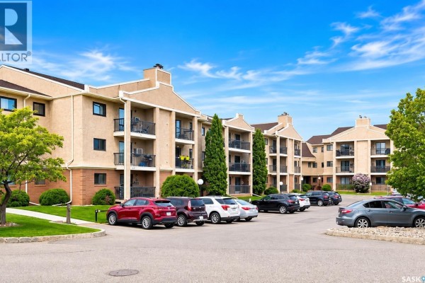 Apartment building exterior with a parking lot in the foreground and trees on the side. - 216 3420 Park Street, Regina
