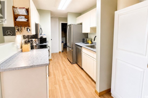Apartment kitchen with light wood floors, white cabinets, stainless steel appliances, and a doorway. - 216 3420 Park Street, Regina