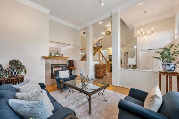 Living room with a stone fireplace, blue upholstered furniture, glass coffee table, and staircase in the background. - 21679 52 Avenue, Langley
