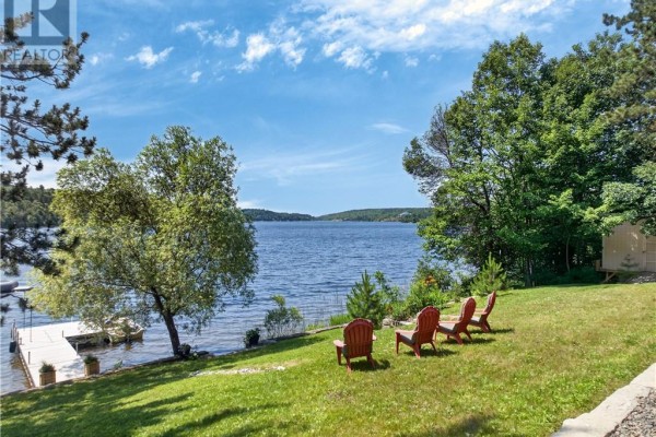 House on a grassy slope with a lake in the foreground, surrounded by trees at sunset. - 2171 Sunnyside Road, Sudbury