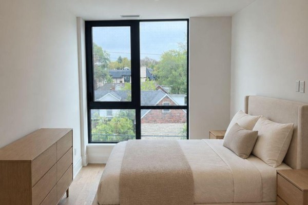 Bedroom with a queen-size bed, nightstands, and dresser beside a large window with a neighborhood view. - 218 - 2788 Bathurst Street, Toronto