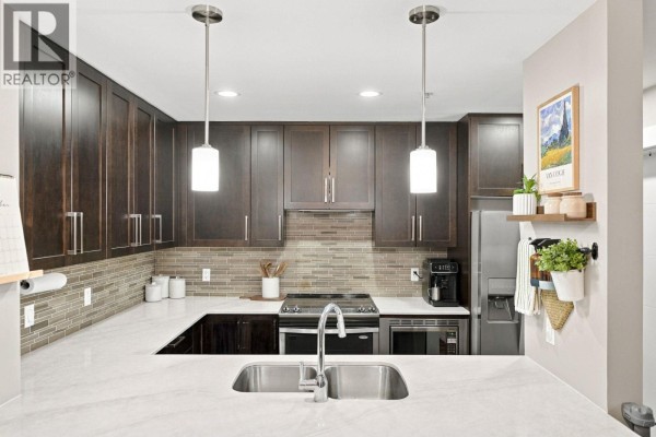 Kitchen island with a double sink, pendant lights, and dark wood cabinetry background. - 218 2495 Wilson Avenue, Port Coquitlam