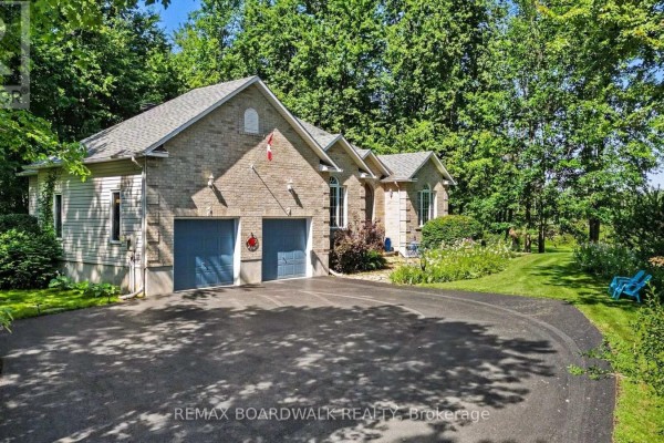 Side view of a light brick home with a two-car garage, featuring a paved driveway and surrounding trees. - 2180 Conley Road, Ottawa