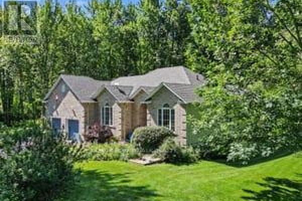 A wide shot of the rear of a brick bungalow nestled amongst tall green trees, with a green lawn. - 2180 Conley Road, Ottawa