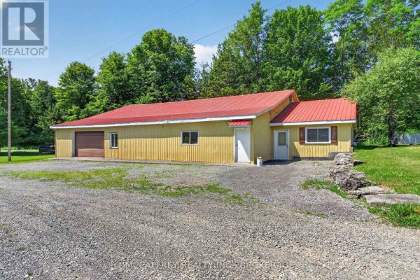 Yellow detached outbuilding with a red metal roof, large garage door, and gravel driveway. - 2183 Daley Road, Tyendinaga