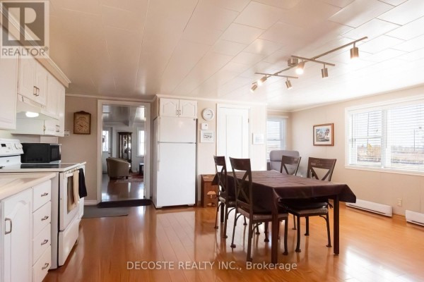 Open-concept dining and living area with white cabinets, a table, and two armchairs near windows. - 21930 Menard Road, North Glengarry