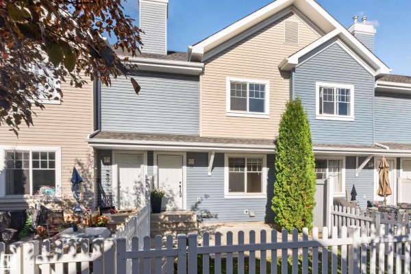 Front exterior of beige, light blue, and gray townhouses with white picket fences and trees. - #22 8304 11 Av Sw, Edmonton