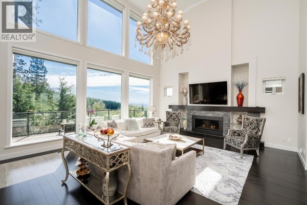 A living room with a fireplace, seating area, and expansive windows with a forest view. - 22 Heritage Peak Road, Port Moody