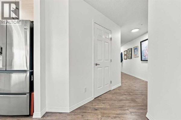 Apartment hallway with white doors, a stainless steel refrigerator, and artwork on the wall. - 220, 2022 Canyon Meadows Drive Se, Calgary