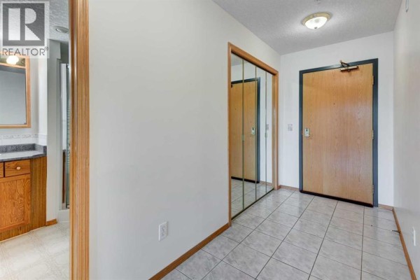 Apartment hallway with tiled floor, mirrored closet doors, and wooden door. - 220, 7229 Sierra Morena Boulevard Sw, Calgary