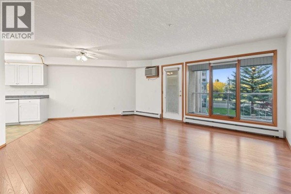 Living room with hardwood floors, white cabinets, and large windows. - 220, 7229 Sierra Morena Boulevard Sw, Calgary